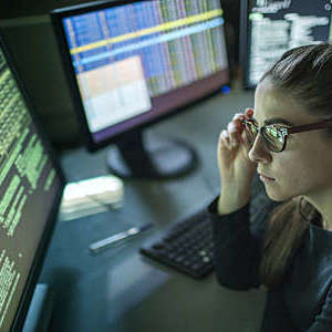 A young woman is seated at a desk surrounded by monitors displaying data, she is contemplating in this dark, moody office.
