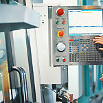 Closeup rear view of a control room at a modern industrial production line with a control person in charge keeping track of the process on a computer. Employee is operating a CNC machine.