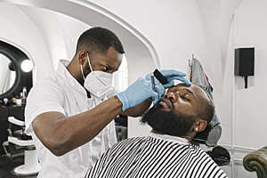 Lithuania, Vilnius, Barber wearing face mask and reusable gloves doing beard of customer in a barber shop