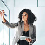 Shot of a young businesswoman using a digital tablet while writing notes on a glass wall in an office
